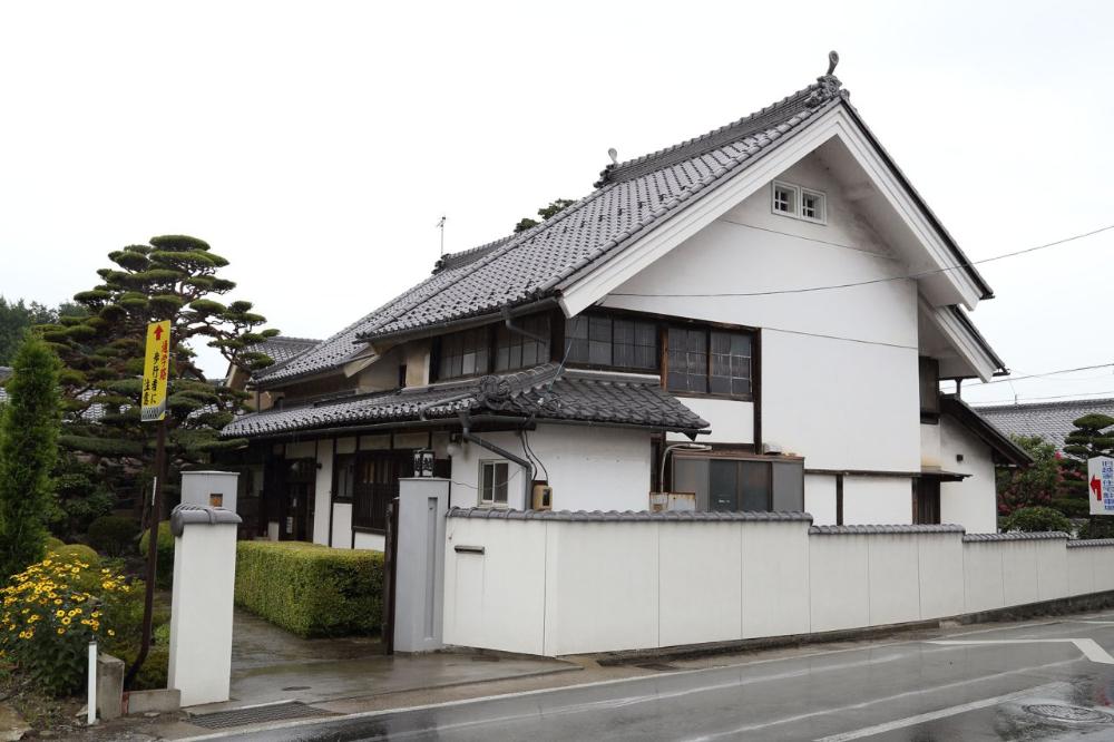 濡れたアスファルトの道路沿いに、白い塀と生け垣に囲まれた、黒い瓦屋根と白い壁が特徴的な入母屋造りの日本家屋が建ち、手前には黄色い花と手入れされた黒松の植栽が見える、曇りの日の街角の風景を捉えた写真。