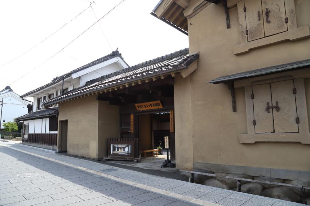 淡い茶色の漆喰壁を持つ蔵造りの建物が、瓦敷きの歩道に沿って並び、右側の建物の二階と一階には黒い金具で留められた重厚な観音開きの土扉が窓として見え、中央の建物の軒下には木製の看板が吊るされ、歴史的な街並みを捉えた写真。