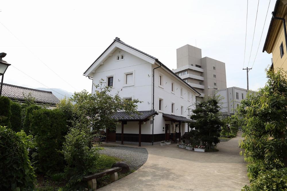 白い壁と黒い瓦屋根が特徴的な伝統的な日本家屋の建物が、手前に植栽と砂利の小道、奥に近代的な高層ビルを背景にしてたたずむ様子を捉えた写真。