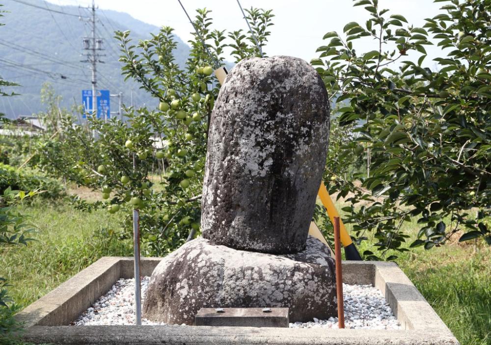 苔と地衣類に覆われた自然石の古い石碑が、コンクリートの低い囲いの中に白っぽい砂利を敷き詰めた台座に立っている写真