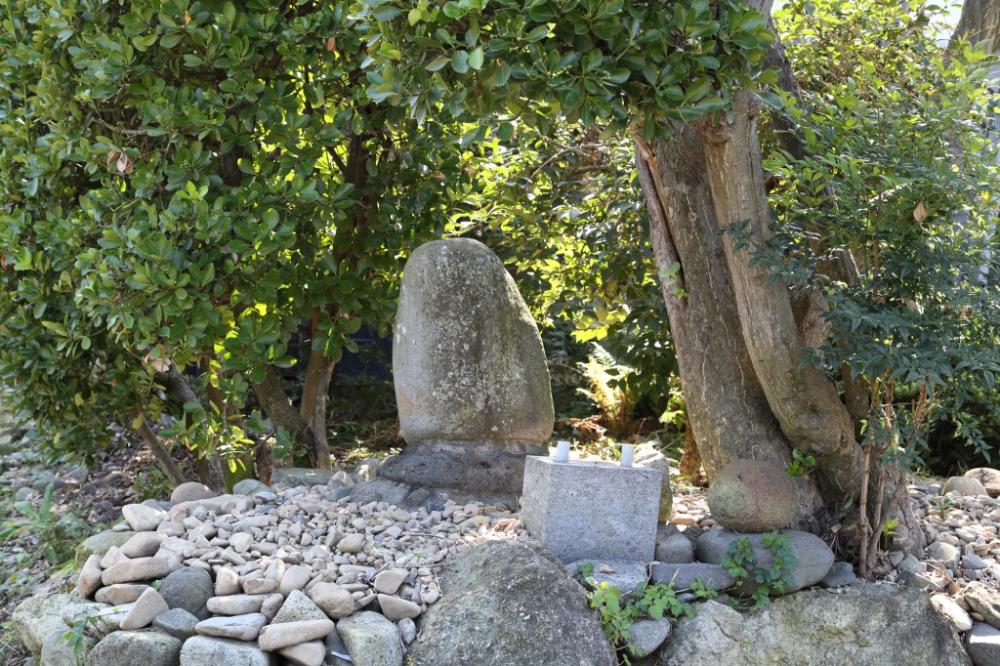 丸みを帯びた縦長の自然石が、木の幹の隣に立っており、その周囲は小石で覆われ、手前には直方体の石の台座のようなものが配置されている虎御前の写真