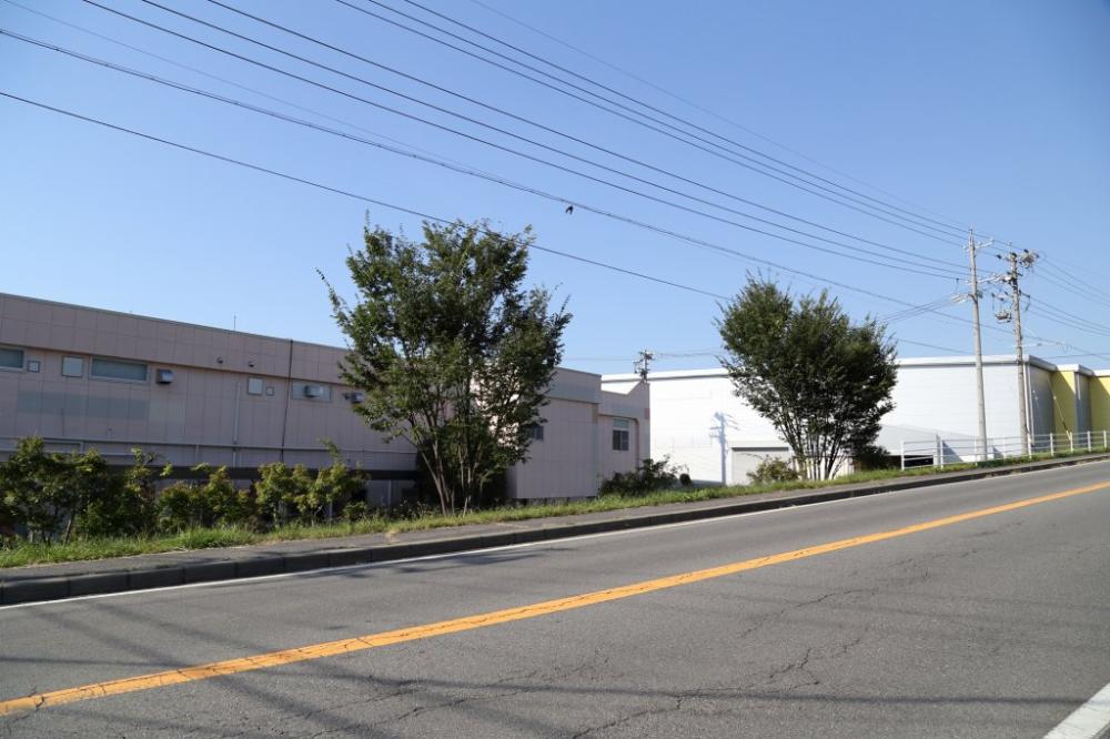 中央に黄色い実線が引かれた道路の右側に、電力線と電柱が沿い、道路の左側には薄いピンク色と白色の大きな建物が並び、その間にいくつかの木々が立っている旧米持神社跡地の写真