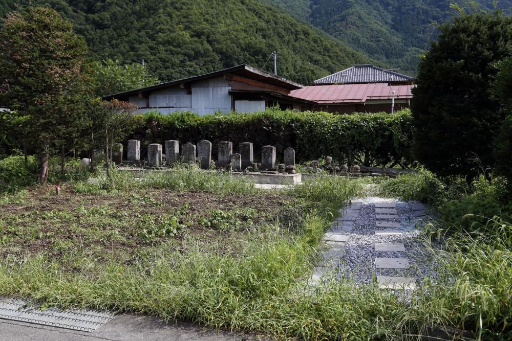 日本家屋が建つ敷地の手前に広がる荒れた庭園の中に、敷石と玉砂利で舗装された通路が伸びており、その奥の低い生け垣の前に、縦長の複数の古い墓石が横一列に並んでいる様子の常楽寺跡写真
