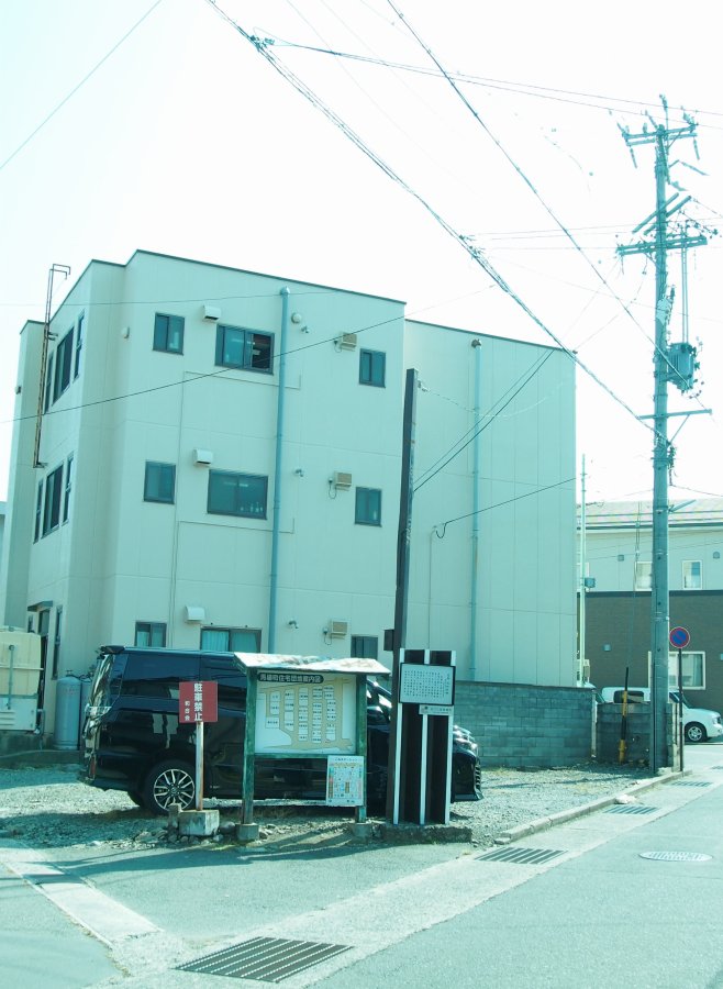 白い三階建ての建物と、その前にある電柱、そして手前の舗装されていない駐車場に駐車されている黒いワンボックスカーと掲示板を撮影した俊明社跡地の写真